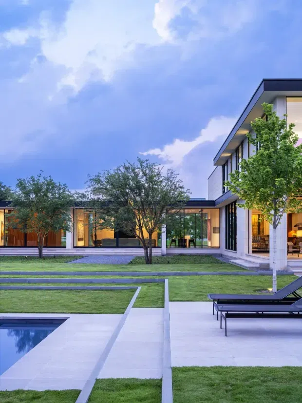 A contemporary courtyard garden viewed from the pool terrace, with a low horizontal wing and taller two-storey glazed volume forming an L-shaped composition. The house features dark flat roof edges, extensive floor-to-ceiling glazing, pale masonry or rendered walls, warm timber interiors, manicured lawns, geometric pale paving, stepped landscaping, and a rectangular swimming pool. Black-framed outdoor loungers sit beside the terrace beneath a dramatic blue evening sky.