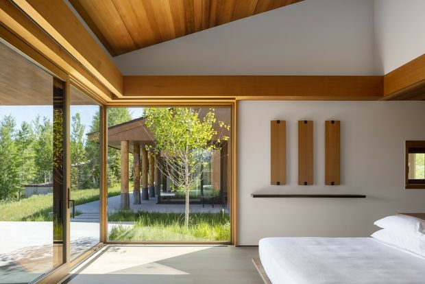 A bright, refined bedroom with large floor-to-ceiling timber-framed glazing opening toward a landscaped courtyard and covered outdoor walkway. The room features a warm timber-lined ceiling, white walls, pale flooring, a simple bed, and minimalist timber wall panels above a slim shelf. The composition emphasizes indoor-outdoor connection, natural light, and clean modern detailing.