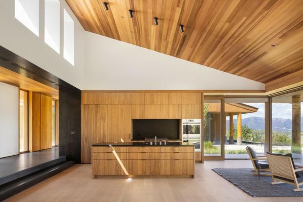 A bright, open-plan contemporary living and kitchen space with extensive warm timber joinery and a dramatic timber-lined ceiling. The kitchen features full-height walnut-toned cabinetry, a dark stone benchtop, integrated appliances, and a central island. A raised entry or circulation platform with timber-framed glazing is visible to the left, while full-height glazing on the right opens to an outdoor terrace and mountain views. The room includes a dining area with woven timber chairs and a dark rug. Clean lines, clerestory windows, and abundant natural light create a refined, warm atmosphere.