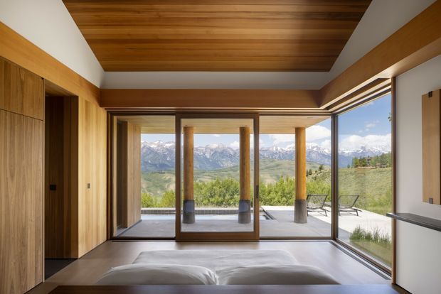 A contemporary bedroom with warm timber wall panelling and a timber-lined ceiling, opening through extensive floor-to-ceiling glazing to a covered terrace and expansive mountain landscape. A large framed glazed opening and built-in timber joinery emphasize the strong indoor–outdoor connection.