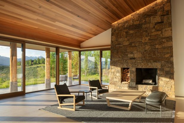 A spacious, light-filled living room with expansive floor-to-ceiling glazing overlooking a landscaped rural setting and distant mountains. The room features a warm timber-lined sloped ceiling, natural stone fireplace wall with a built-in hearth, minimalist lounge chairs, a low coffee table, and a large neutral rug. Strong indoor-outdoor connection and contemporary lodge-like character.