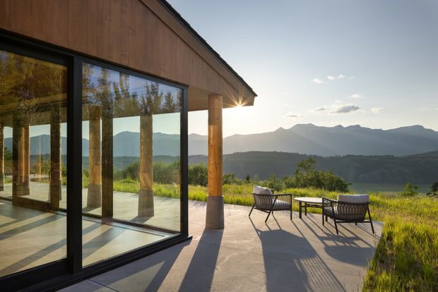 A contemporary timber-clad pavilion or house extension with extensive floor-to-ceiling glazing opens onto a broad concrete terrace. Timber structural posts and dark-framed sliding glass doors create a strong indoor-outdoor connection, with simple outdoor seating overlooking a dramatic mountain and valley landscape in warm late-afternoon light.