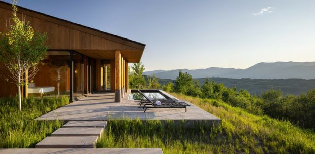 A contemporary single-storey house set on a sloping grassy site, with a broad low-pitched roof, warm timber-clad facade, extensive glazing, and a projecting paved terrace furnished with lounge chairs. A stepping-stone path approaches the terrace, while layered mountain ranges form the distant backdrop.