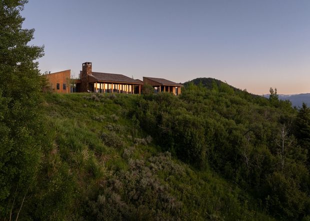 A low-profile mountain residence set along a heavily vegetated hillside at dusk. The elongated building has warm timber-toned exterior walls, extensive vertical glazing, dark pitched roofs, and a prominent stone chimney. Forested terrain dominates the foreground, with distant mountain or valley views visible at the right.