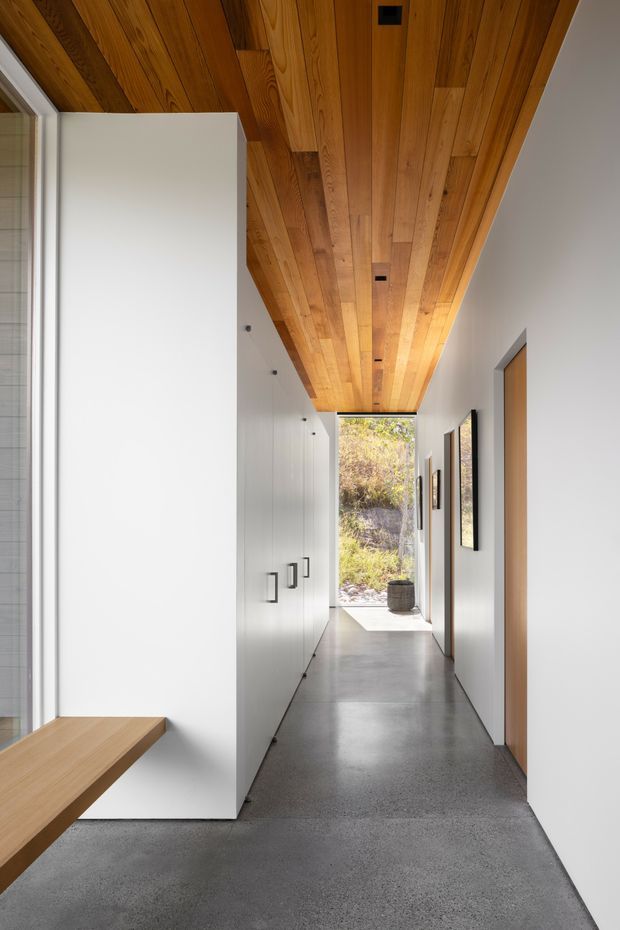 A long, narrow minimalist hallway with a polished grey concrete floor, white built-in cabinetry and flush doors, and a striking warm timber-lined ceiling with recessed downlights. Framed artwork lines the right wall, while the corridor opens to a sunlit planted outdoor area at the far end. A partial timber bench is visible in the foreground.
