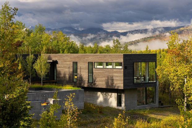 A modern two-level home set within a wooded, mountainous landscape. The building has a long, rectilinear form with dark charcoal cladding, warm horizontal timber-look cladding, extensive floor-to-ceiling glazing, and a partially recessed lower level. A concrete outdoor terrace or fire feature sits beside the house, while autumn vegetation and misty mountain ridges surround the property.