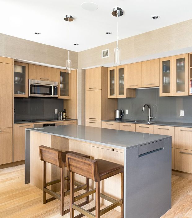 Bright contemporary kitchen with full-height light timber cabinetry, integrated appliances, dark grey splashback and benchtops, and a large central island with seating for three. The space has a clean, streamlined fit-out, pendant lighting, timber flooring, and neutral textured walls.