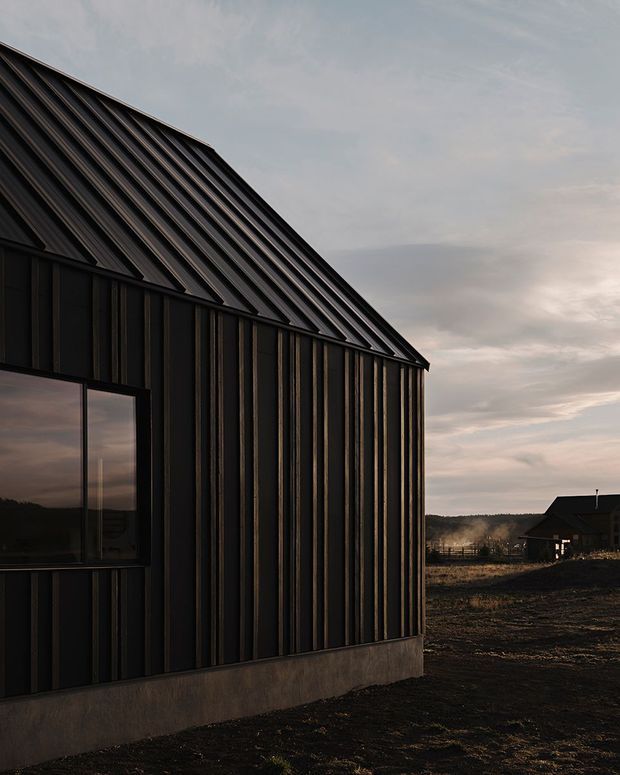 Exterior view of a dark, barn-like building with a steep pitched gable roof and vertically ribbed metal cladding. A large reflective window is set into the side facade, while a rural landscape and another outbuilding sit in the background under an overcast sky.
