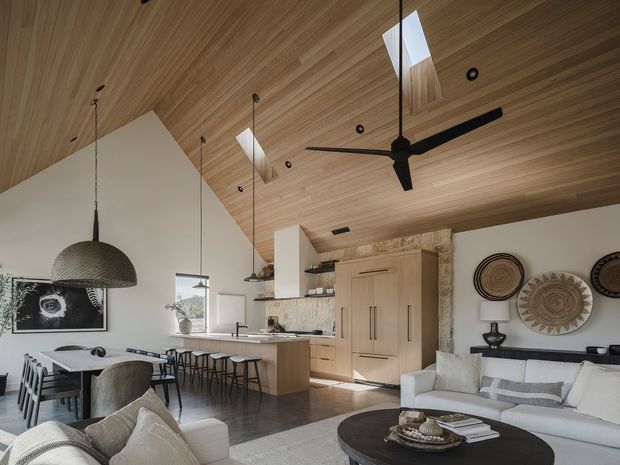 Open-plan contemporary living, dining and kitchen area with a warm timber-lined ceiling, exposed stone wall, large kitchen island, dining table, neutral upholstered seating, black pendant lights, and a large ceiling fan. Skylights and clerestory-style openings bring natural light into the space.