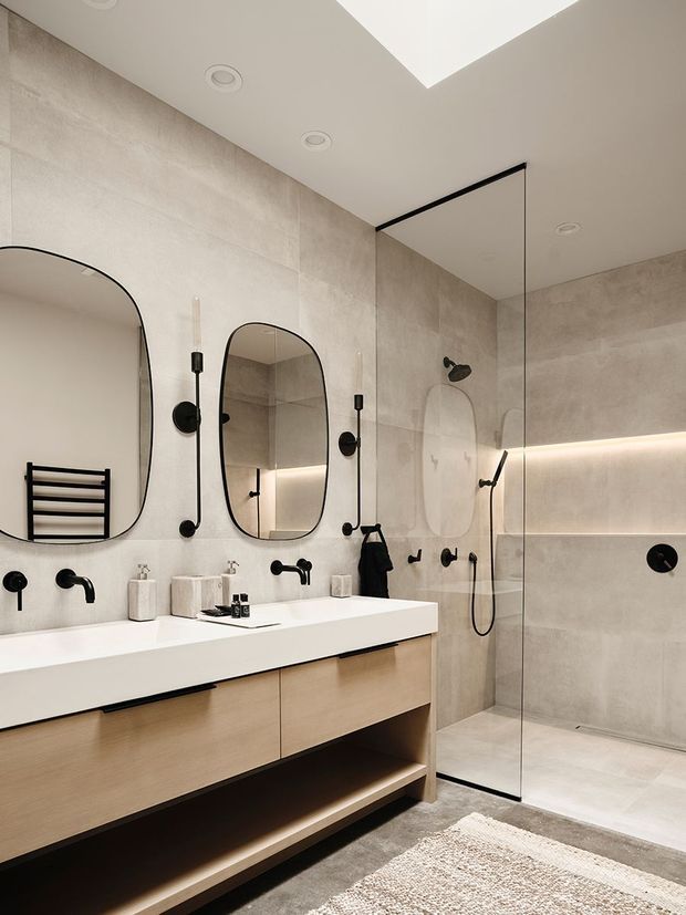 Contemporary bathroom with a long floating timber vanity, white integrated benchtop, twin wall-mounted black taps and basins, rounded-rectangle mirrors, a black heated towel rail, and a frameless glass walk-in shower. Large pale beige-grey tiled walls and floor are complemented by recessed ceiling lights and concealed warm vanity/shower lighting.