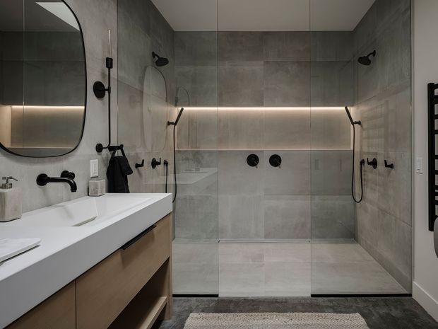 Contemporary bathroom featuring a large walk-in glass shower with dual black shower fittings, full-height grey stone-look wall and floor tiles, a floating timber vanity with white integrated basin, black tapware, rounded-edge mirror, and concealed horizontal LED lighting.
