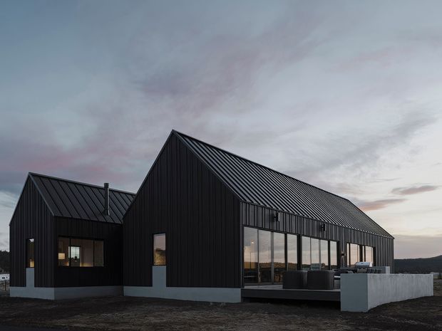 Exterior view of a contemporary dark-clad house composed of intersecting pitched-roof volumes. The building has black vertical cladding, ribbed metal roofing, large glazed openings, and a sheltered outdoor terrace with a low concrete wall. It is photographed at dusk against an open landscape and cloudy sky.