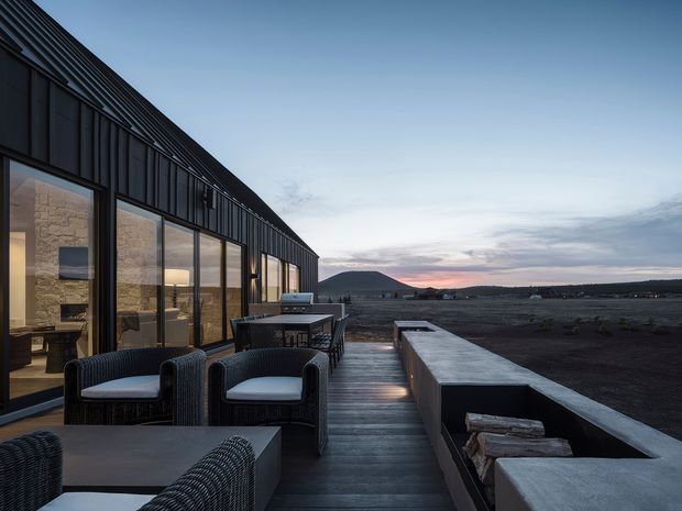A contemporary dark-clad building with a long sloping roof and extensive floor-to-ceiling glazing, viewed from a broad timber deck at dusk. The terrace includes woven outdoor lounge chairs, a low table, built-in concrete seating or planter walls, and a linear outdoor fireplace feature containing timber logs. Warm interior light reveals stone walls and furnishings inside, while the surrounding open landscape and distant hills form the backdrop.