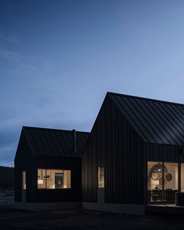 A dark, modern gabled building photographed at dusk. The exterior is clad in vertically ribbed black metal siding with matching pitched standing-seam metal roofs. Warm interior lighting glows through large windows and reveals furnished rooms inside, while a forested landscape sits in the distant background.