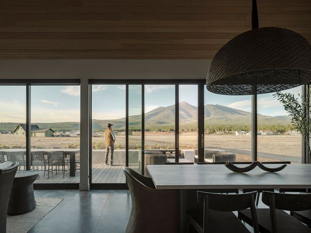 A contemporary dining and living area with expansive floor-to-ceiling glazing and sliding doors opening to an outdoor terrace. The room features a long dining table, dark woven dining chairs, a large woven pendant light, polished concrete flooring, and a timber-lined ceiling. Mountain and rural scenery are visible beyond the glazing.