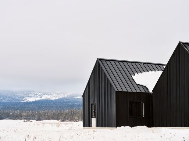 A dark, modern gabled building clad in vertically ribbed black metal stands in a snow-covered rural landscape. The composition shows two connected pitched-roof volumes, heavy snow on the roof and ground, and a forested mountain backdrop beneath an overcast sky.