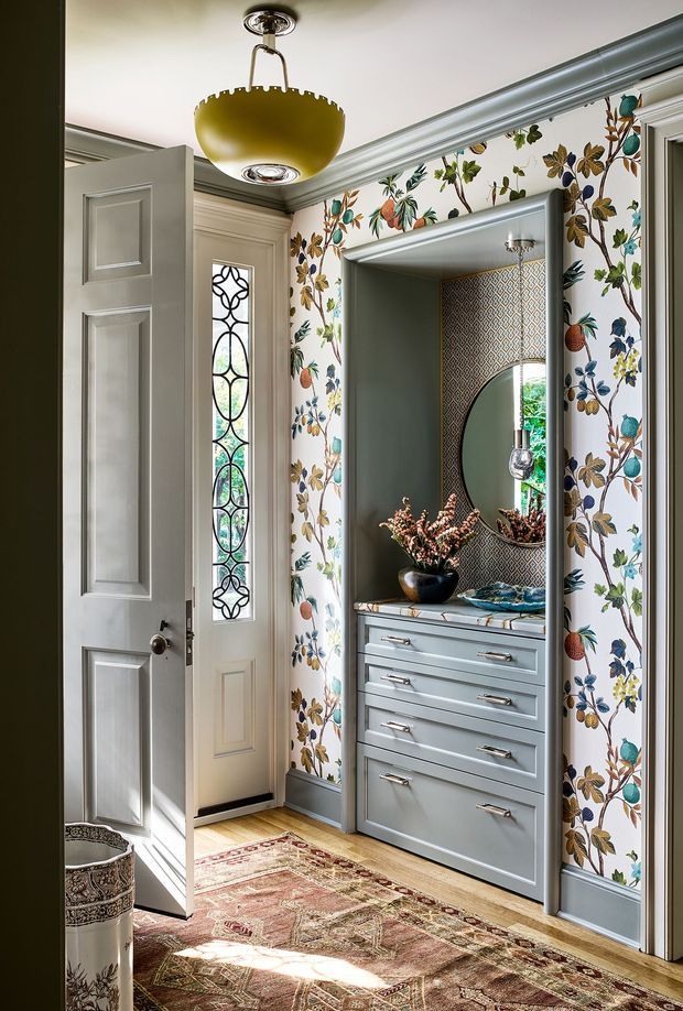 A richly detailed traditional entryway with pale grey-blue panelled doors, ornate leaded-glass glazing, botanical patterned wallpaper, and a built-in grey-blue dresser with multiple drawers. The recessed niche includes patterned tile or wallpaper, a round mirror, decorative lighting, and floral arrangements. A patterned vintage-style rug covers the timber floor, while a brass-toned pendant light and layered mouldings add character.