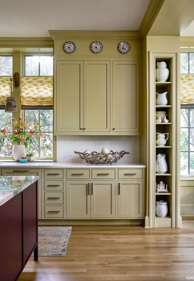 A traditional-style kitchen with muted olive-beige built-in cabinetry, framed panel doors, brass-toned hardware, and a white benchtop. A dark burgundy island with a pale stone or marble surface occupies the foreground. The room features decorative cornice and trim, patterned botanical window coverings, large windows, timber flooring, and a tall open display niche filled with white ceramic vessels. Decorative plates and a sculptural bowl add visual interest.