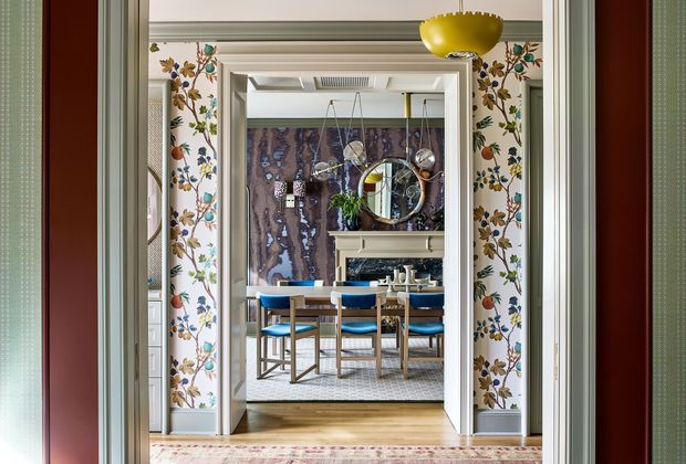 A richly layered interior view through aligned doorways, showing a formal dining room beyond a hallway or entry passage. The walls feature ornate floral wallpaper framed by painted architectural mouldings and trim. A round dining table with upholstered blue chairs sits on a pale rug, with a dark dramatic marble or stone fireplace wall and reflective decorative elements behind it. A sculptural yellow pendant light hangs in the foreground, while timber flooring, layered rugs, and detailed traditional joinery create a refined, colourful atmosphere.