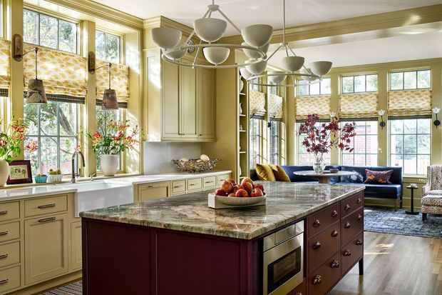 A richly detailed traditional kitchen with a large central island, inset cabinetry, marble benchtops, integrated cooking appliances, and a casual living area beyond. Tall multi-pane windows feature patterned Roman blinds, while layered pendant lighting, decorative trim, and warm olive-green cabinetry create an elegant, heritage-inspired interior.