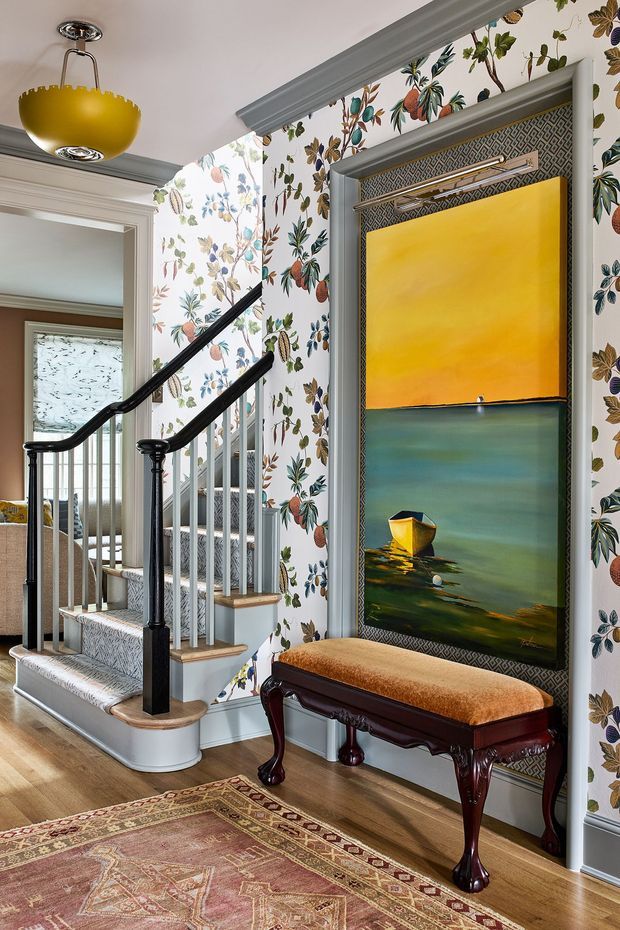 A richly decorated residential stairway and landing with a painted timber staircase, white balusters, black handrails and posts, ornate floral wallpaper, grey architectural trim, and warm timber flooring. A framed colourful artwork is mounted above a traditional upholstered bench, with a patterned area rug in the foreground. The space has layered traditional detailing and an eclectic, colourful interior character.