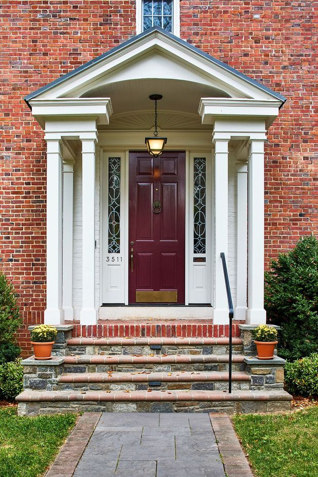 Front exterior entrance of a traditional brick residence, featuring a formal covered portico with white classical columns, pilasters, moulded trim, and a triangular pediment. A dark burgundy paneled door is flanked by decorative leaded-glass sidelights, with brick and stone steps leading up from a paved garden path. Potted plants and clipped shrubs frame the entry.
