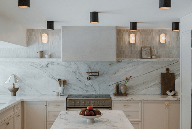 Refined contemporary kitchen with pale cabinetry, a large veined marble backsplash and benchtops, a central island, and a freestanding-style range integrated into the cabinetry. Warm pendant and wall lighting contrasts with the cool stone surfaces, while timber boards and restrained styling add warmth.