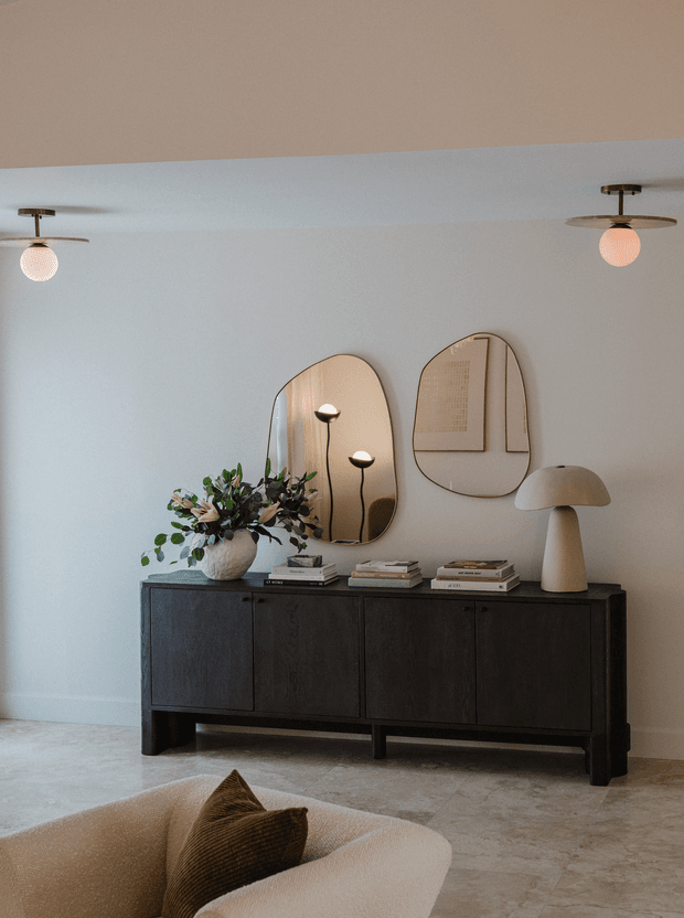 A refined living room vignette featuring a dark wood sideboard, sculptural neutral table lamp, floral arrangement, stacked books, organic-shaped mirrors, framed artwork, and globe ceiling lights. A curved upholstered sofa with a textured cushion is visible in the foreground. The palette is warm and neutral with strong black-brown joinery contrast.