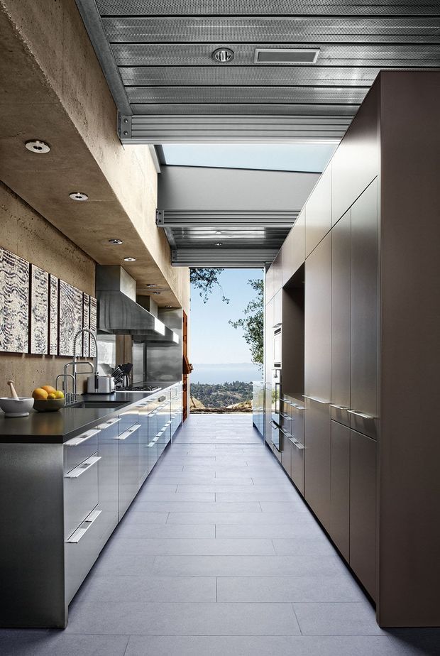 A narrow, elongated contemporary galley kitchen with full-height dark grey cabinetry on both sides, integrated appliances, a long dark benchtop, stainless steel sink and cooktop, and a central tiled walkway. The ceiling combines exposed concrete with extensive ribbed metal panels and recessed spotlights, with a large skylight or glazed opening leading toward a distant outdoor view. The space has a refined industrial character and strong linear perspective.