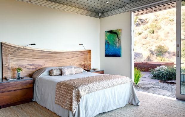 A bright, contemporary bedroom with a low-profile bed dressed in white and taupe layered bedding, a sculptural timber headboard, dark timber bedside cabinetry, and slim reading lights. Full-height glazed doors open to a landscaped exterior, while a colourful abstract artwork and timber-lined ceiling add visual interest.