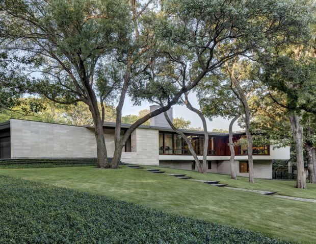 A low-profile modern residence set within a mature, carefully landscaped garden. The facade combines pale horizontal masonry or stonework with dark glazing, deep roof planes, and a recessed upper-level section featuring warm timber-toned cladding. Large established trees frame the house, while clipped groundcover, lawn, and stepping-stone paths create a restrained, integrated landscape.