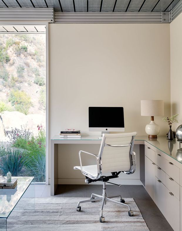 A bright, compact home office with a built-in L-shaped desk and drawers, glass desktop surfaces, a large desktop computer, and a modern white ergonomic office chair. A floor-to-ceiling glazed opening provides views of landscaped terrain, while a metal-lined ceiling and pale neutral walls create a clean contemporary setting. A table lamp, books, vase, and rug add restrained decorative detail.