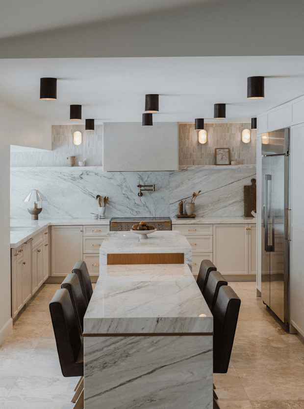 A refined kitchen and dining space with a large marble-clad island extending into a dining table, surrounded by dark upholstered chairs. The kitchen features pale painted cabinetry with brass-toned hardware, a dramatic continuous veined marble splashback and benchtops, integrated stainless-steel appliances, and a warm textured tile backsplash. Dark cylindrical pendant and wall lights contrast with the light surfaces, creating a sophisticated, contemporary interior.