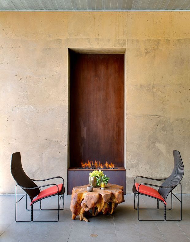 A refined interior vignette with a tall recessed dark timber fireplace surround set into a warm, textured plaster or concrete wall. A linear flame burns above a dark built-in hearth cabinet, flanked symmetrically by two black woven lounge chairs with vivid red cushions. A sculptural natural timber stump coffee table with a small plant sits in the foreground. The space has a high pale timber-lined ceiling and a restrained, earthy palette with bold red accents.