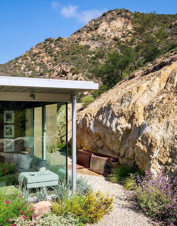 A modern glass-fronted residence set directly against a rugged rocky hillside. The image shows a low, flat-roofed extension with expansive glazing, pale metal roof edging, and a visible lounge interior. A gravel path, built-in seating, and drought-tolerant landscaping occupy the foreground beside the exposed rock face.
