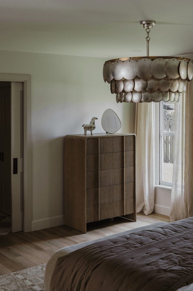A softly lit bedroom with a warm, refined interior. The room features a dark wood bed with rumpled taupe bedding, pale timber flooring, off-white walls, sheer full-length curtains, and a tall fluted wood-front dresser. A sculptural layered-disc pendant light hangs from the ceiling, while a small horse sculpture and round mirror rest on the dresser. A doorway opens to the left.