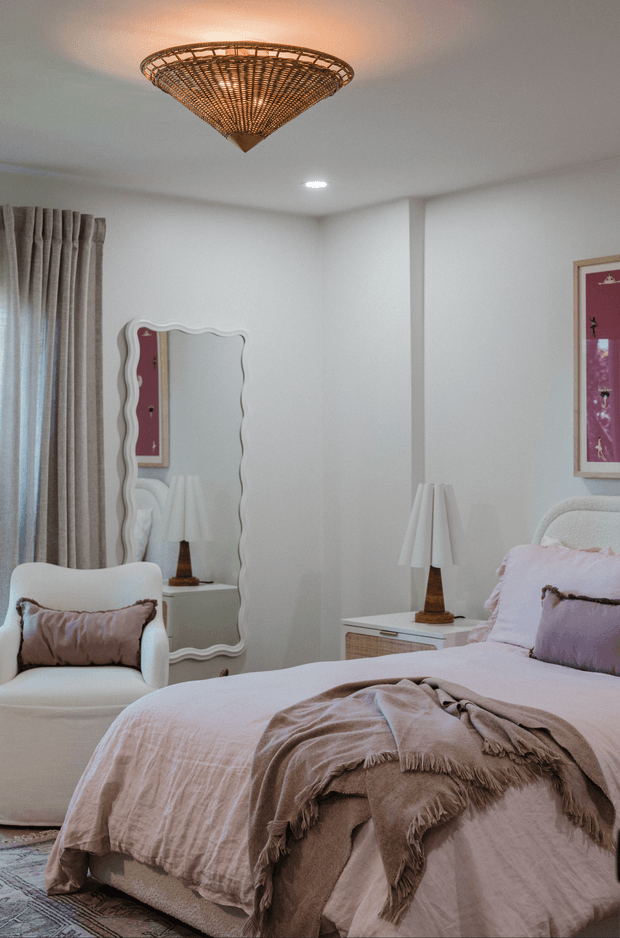 A softly styled bedroom with a large upholstered bed dressed in pale blush bedding and layered taupe throws, a white boucle armchair, bedside tables with pleated lamps, full-length wavy-edged mirror, framed artwork, textured curtains, and a woven statement ceiling light. The palette is warm and muted with refined, feminine detailing.