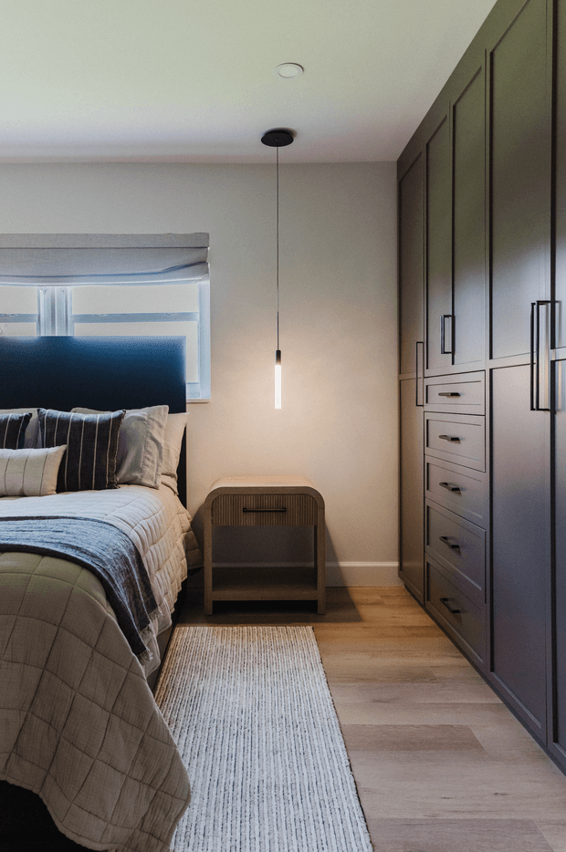 A compact, contemporary bedroom with a partially visible bed dressed in layered neutral bedding, a dark upholstered headboard, a large built-in wall of dark cabinetry, and warm timber flooring. A slim pendant light hangs beside a small wooden bedside table, while a textured light rug runs along the bed. The room has a calm, tailored, softly lit atmosphere with restrained detailing and strong vertical cabinetry lines.