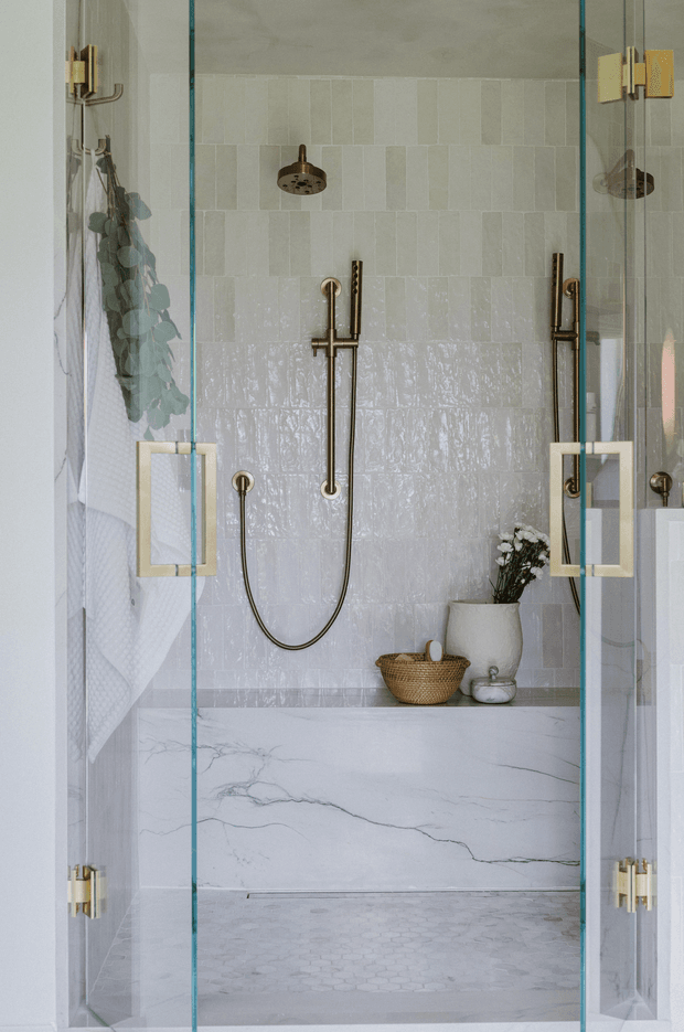 A refined walk-in shower enclosure viewed straight on through clear glass doors. The bathroom features pale textured rectangular wall tiles, a lower band of glossy handmade-look tiles, a marble-look shower bench and wall, small hexagonal floor tiles, and brushed brass shower fittings and hardware. A woven basket, ceramic vessel, and greenery add subtle natural accents.