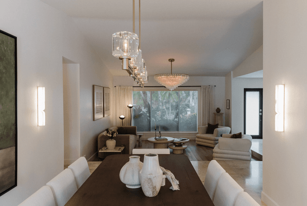 A refined open-plan dining and living area with a dark timber dining table, upholstered white dining chairs, sculptural ceramic vessels, layered pendant lighting, neutral furnishings, and full-width glazing overlooking greenery. The room features white walls, timber flooring, curtains, contemporary artwork, and a softly lit, polished interior.