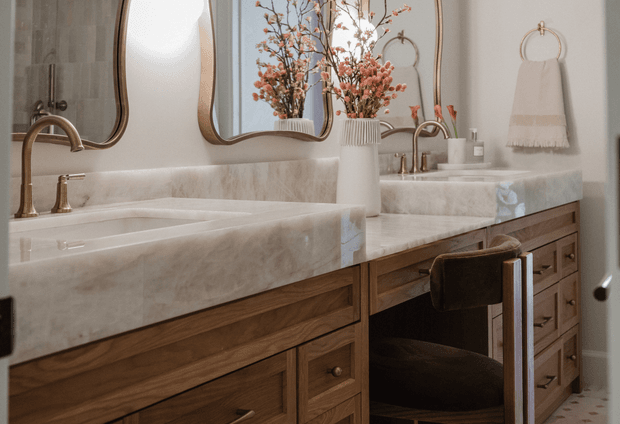 Warm, refined bathroom interior featuring a double vanity with white marble surfaces, timber cabinetry, brass fixtures, rounded mirrors, and a freestanding marble bathtub. A stool is tucked beneath the vanity, while floral branches and textured towels add soft decorative accents.