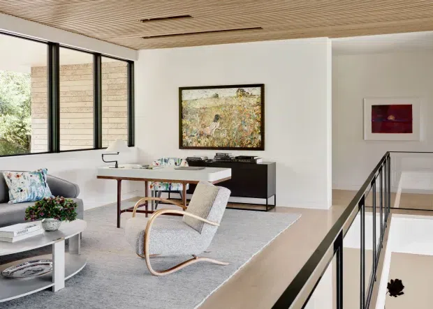 Bright contemporary living room viewed from an upper-level landing, featuring large black-framed windows, a white-painted interior, a slatted timber ceiling with recessed linear lighting, pale timber flooring, a grey area rug, a compact desk, a dark low sideboard, and a sculptural light-toned rocking chair. A black metal balustrade borders the stair opening, with artwork and greenery adding visual interest.
