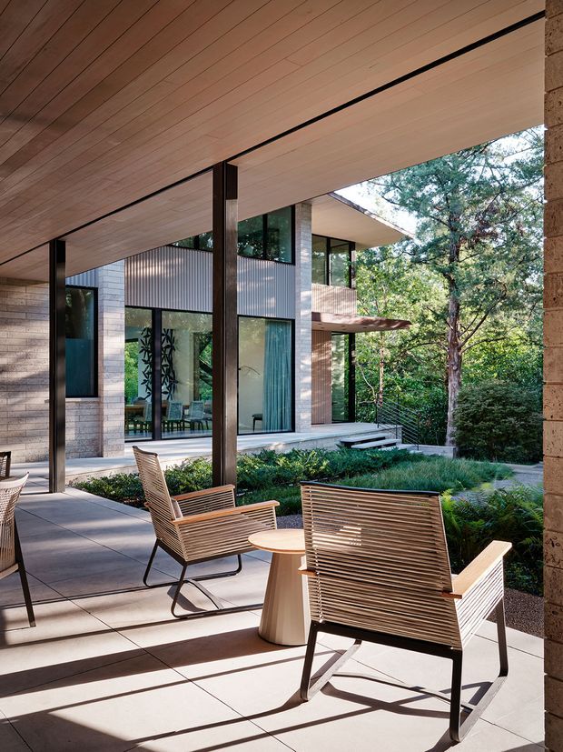 A covered outdoor terrace with timber-lined soffits, slim dark structural posts, concrete paving, and contemporary slatted outdoor lounge chairs surrounding a small round table. The terrace overlooks a landscaped garden and faces a modern house with extensive glazing, pale masonry walls, corrugated metal cladding, and broad horizontal roof planes.