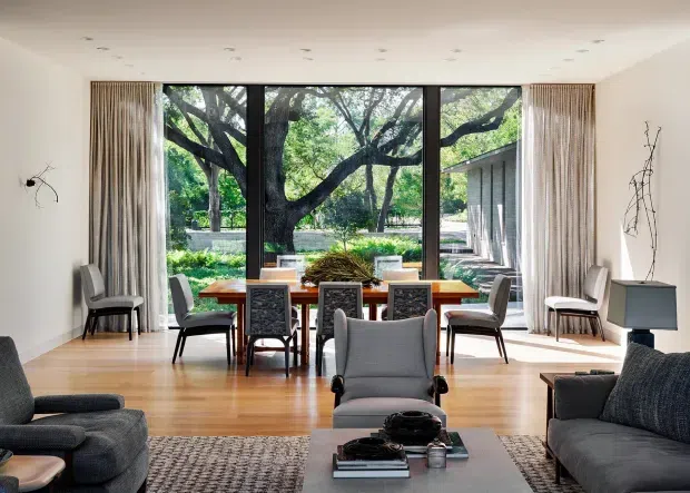 Bright, refined open-plan living and dining area with a contemporary mid-century-inspired interior. A timber dining table and upholstered chairs sit beside full-height black-framed glazing overlooking a leafy garden, while grey sofas and armchairs define the foreground living zone. Pale walls, timber flooring, recessed ceiling lights, and neutral textiles create a calm, airy atmosphere.