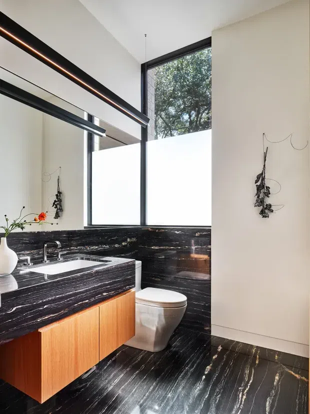 A contemporary bathroom with a wall-mounted timber vanity, white basin and chrome tapware, toilet, and extensive dramatic black marble or natural stone surfaces with strong white veining across the benchtop, splashback, walls, and floor. Large black-framed windows provide abundant natural light, while linear suspended lighting and simple wall-mounted botanical decor add a refined modern character.