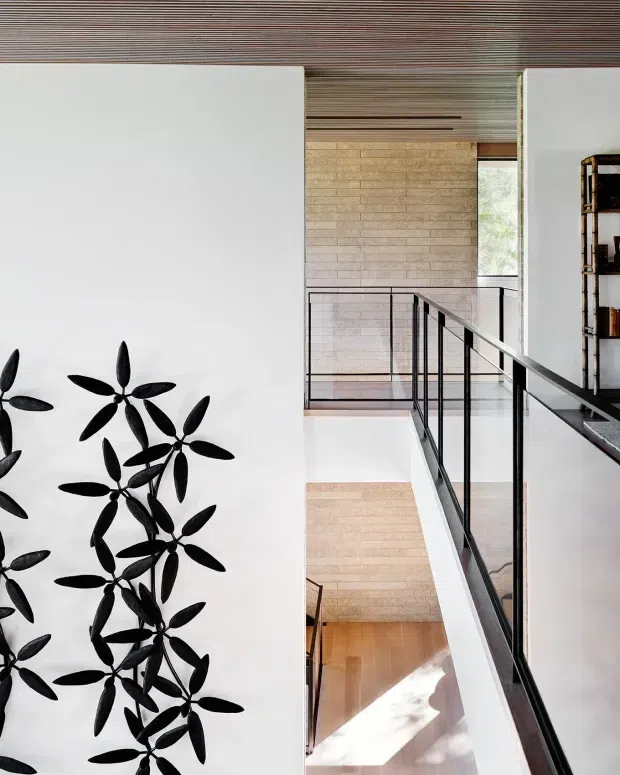 Interior architectural view of a multi-level stairway and landing with slim black metal balustrades, pale timber flooring, light brick or masonry wall surfaces, white walls, and a slatted timber ceiling. A black botanical wall artwork and open shelving are visible at the edges of the frame. Natural light enters from a narrow window, creating a refined, modern vertical circulation space.