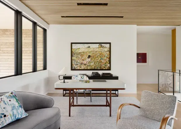 A bright, refined contemporary living room with a grey sofa and armchair, a central timber-legged coffee table, a low black media console, framed artwork, expansive black-framed windows, and a glass stair balustrade. The room features a white wall palette, light grey carpet, and a warm slatted timber ceiling with integrated linear lighting.
