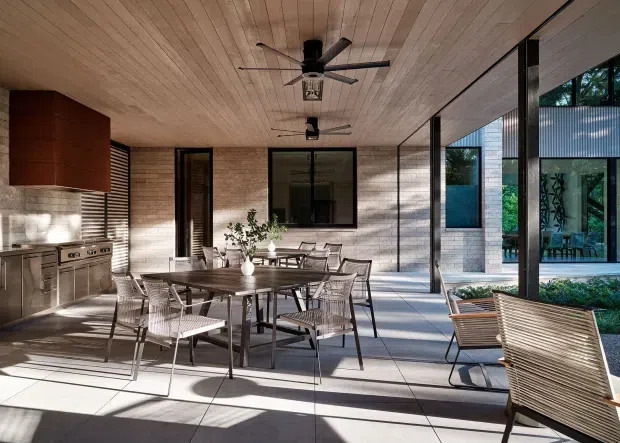 A covered outdoor dining and kitchen area with a contemporary architectural design. The space features a built-in stainless-steel kitchen along a brick wall, a large timber-lined ceiling with multiple ceiling fans, floor-to-ceiling black-framed glazing, and a dining table surrounded by woven chairs. Large pale tiled paving extends through the open-sided room, connecting visually to a landscaped courtyard and adjacent glazed pavilion.