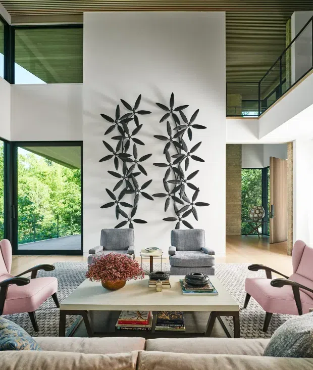 A sophisticated double-height living room with a large white feature wall decorated with sculptural black botanical wall art. Two grey lounge chairs face a low rectangular coffee table, surrounded by pink and neutral upholstered seating. Full-height glazing opens toward lush greenery, while a mezzanine-level walkway and dark slatted ceiling add architectural interest. The palette is refined and softly contrasting, with warm timber and stone details visible beyond the room.