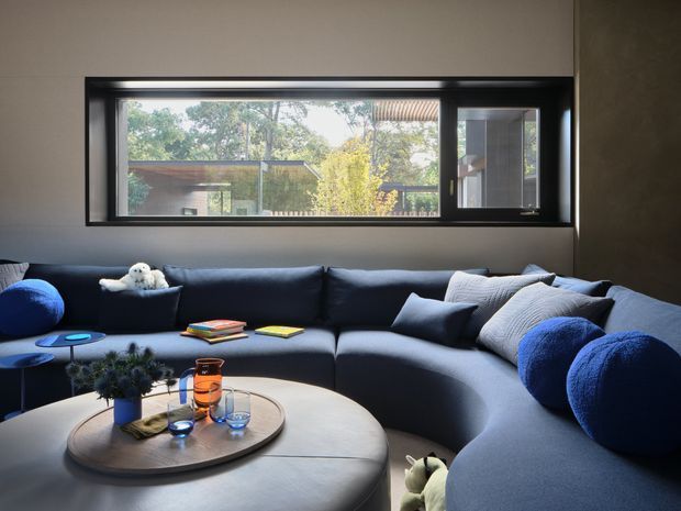 A modern living room centered on a large deep-blue sectional sofa and rounded upholstered coffee table. The room has dark neutral walls, a black-framed horizontal picture window overlooking trees and a neighbouring structure, and a low blue side table. Cushions, books, a small plant, and glassware add restrained decorative detail. The space feels contemporary, comfortable, and slightly moody, with strong natural light entering through the window.
